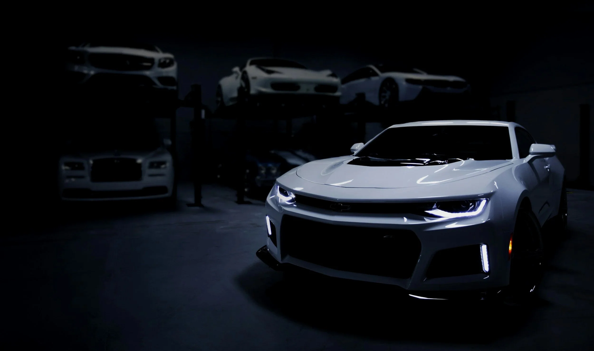 A white sports car with illuminated headlights parked in a dimly lit garage, with other cars visible in the background.