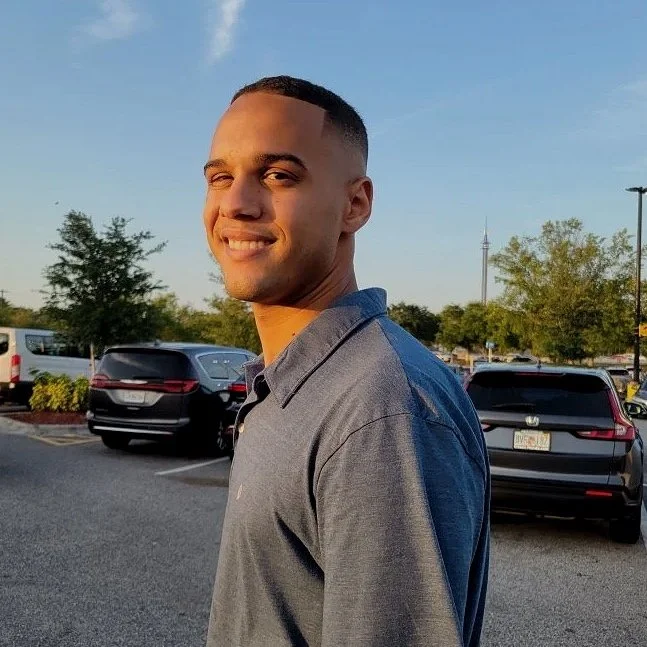 Young man with short hair smiling, standing in a parking lot during sunset, with cars and trees in the background.