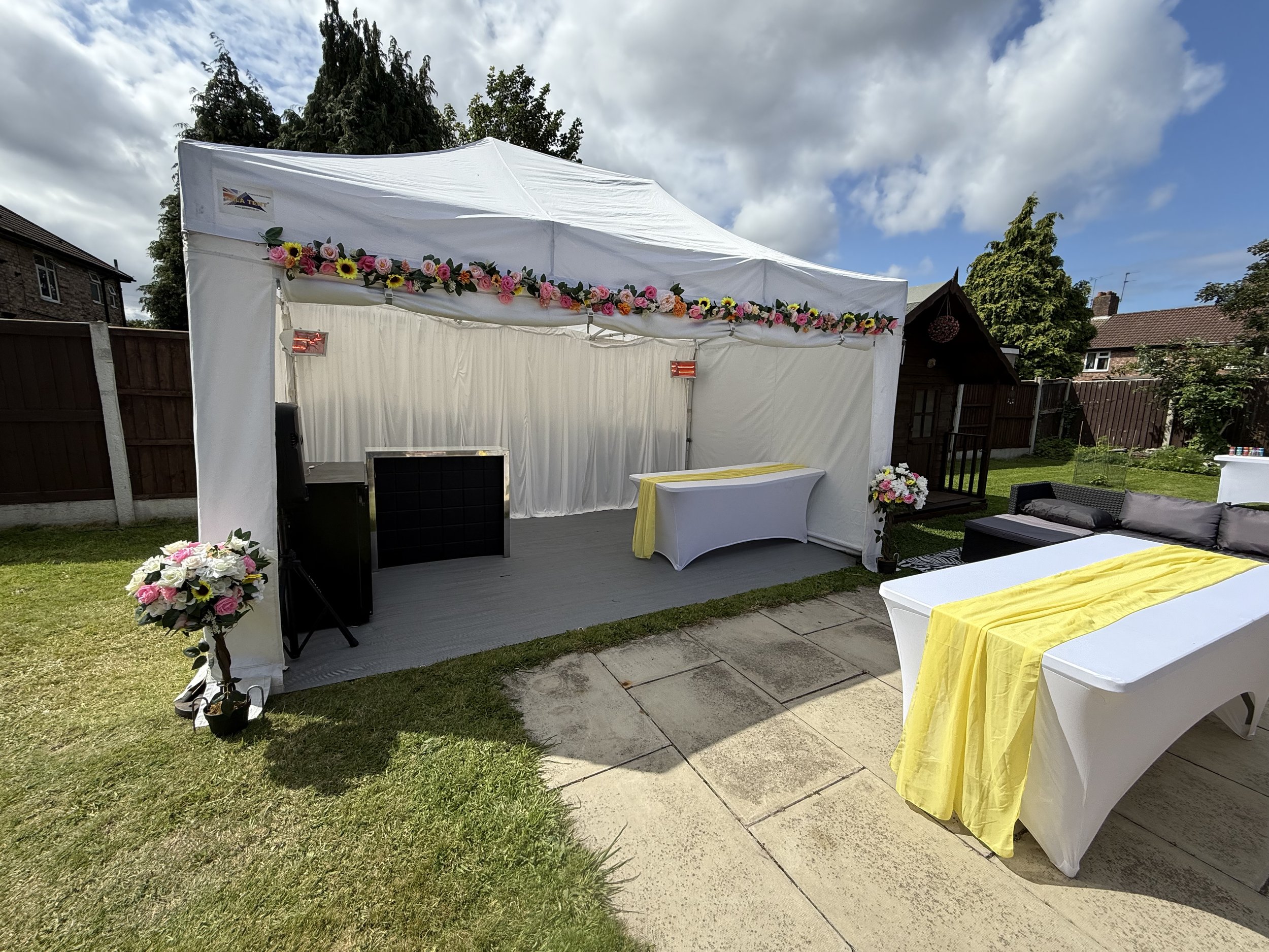 Outdoor event setup with a white tent decorated with pink and yellow flowers, tables covered with white and yellow tablecloths, and floral arrangements on the grass, with a garden and houses in the background under a partly cloudy sky.