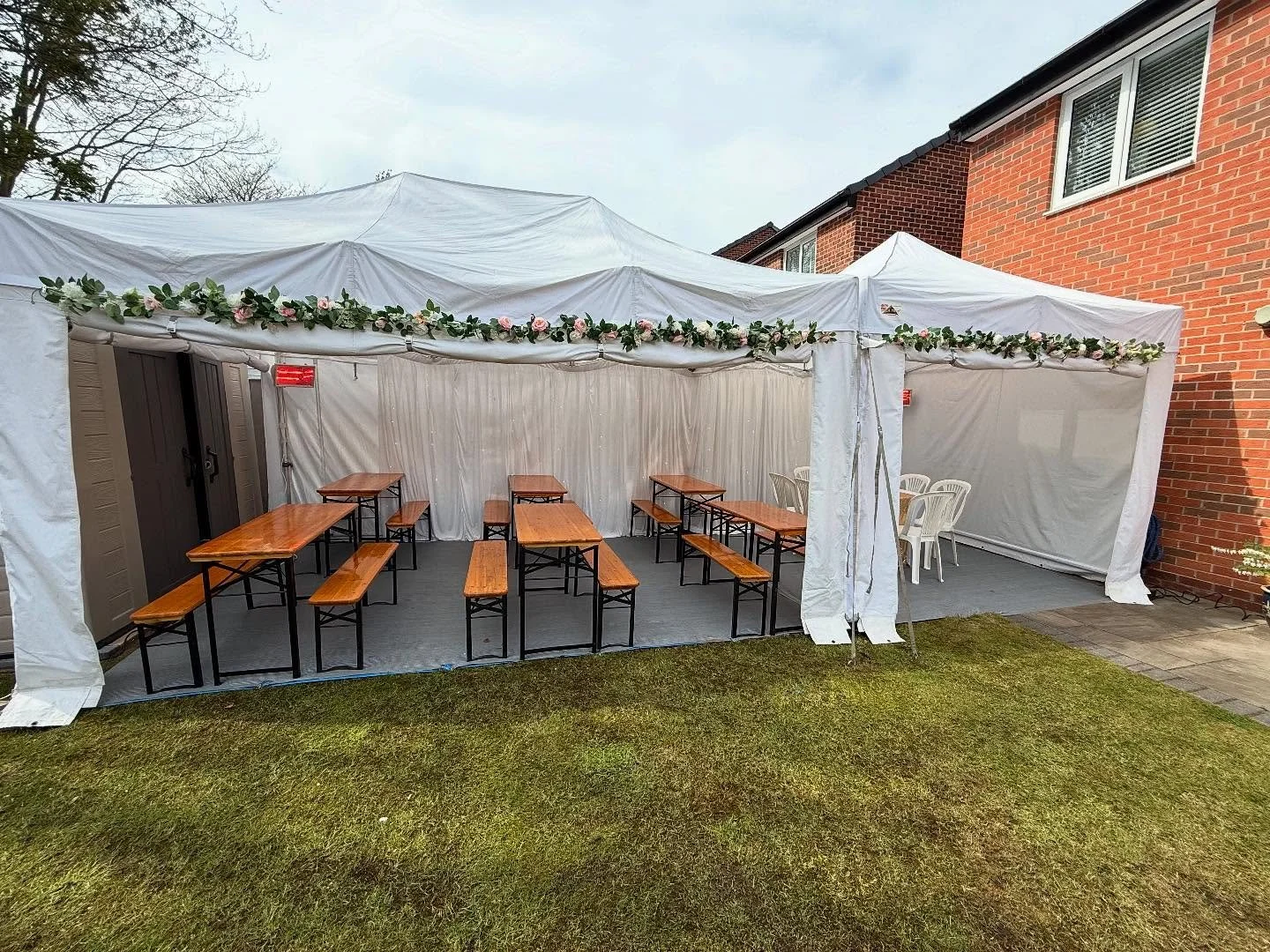 White outdoor event tent with floral garland decoration, wooden tables and benches, and white plastic chairs inside, set up on a grassy backyard next to a brick house.