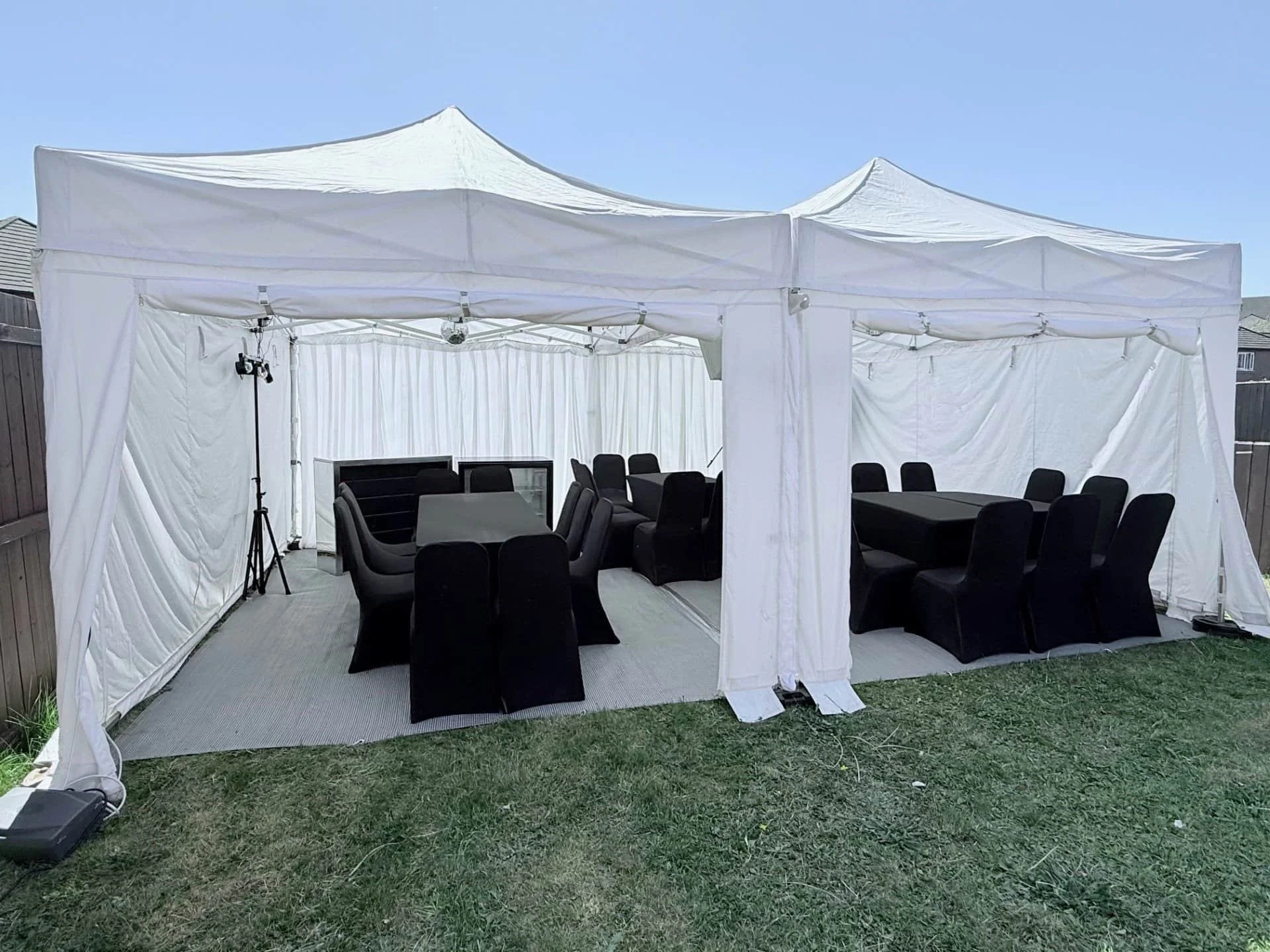 A white outdoor event tent with black chairs and tables inside, set up on a grassy yard.