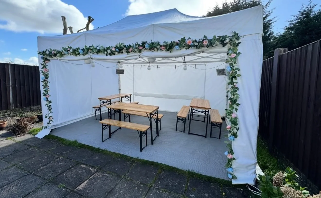 A white outdoor event tent decorated with a floral garland, set up in a backyard with wooden picnic tables and benches inside, under a blue sky with some clouds.