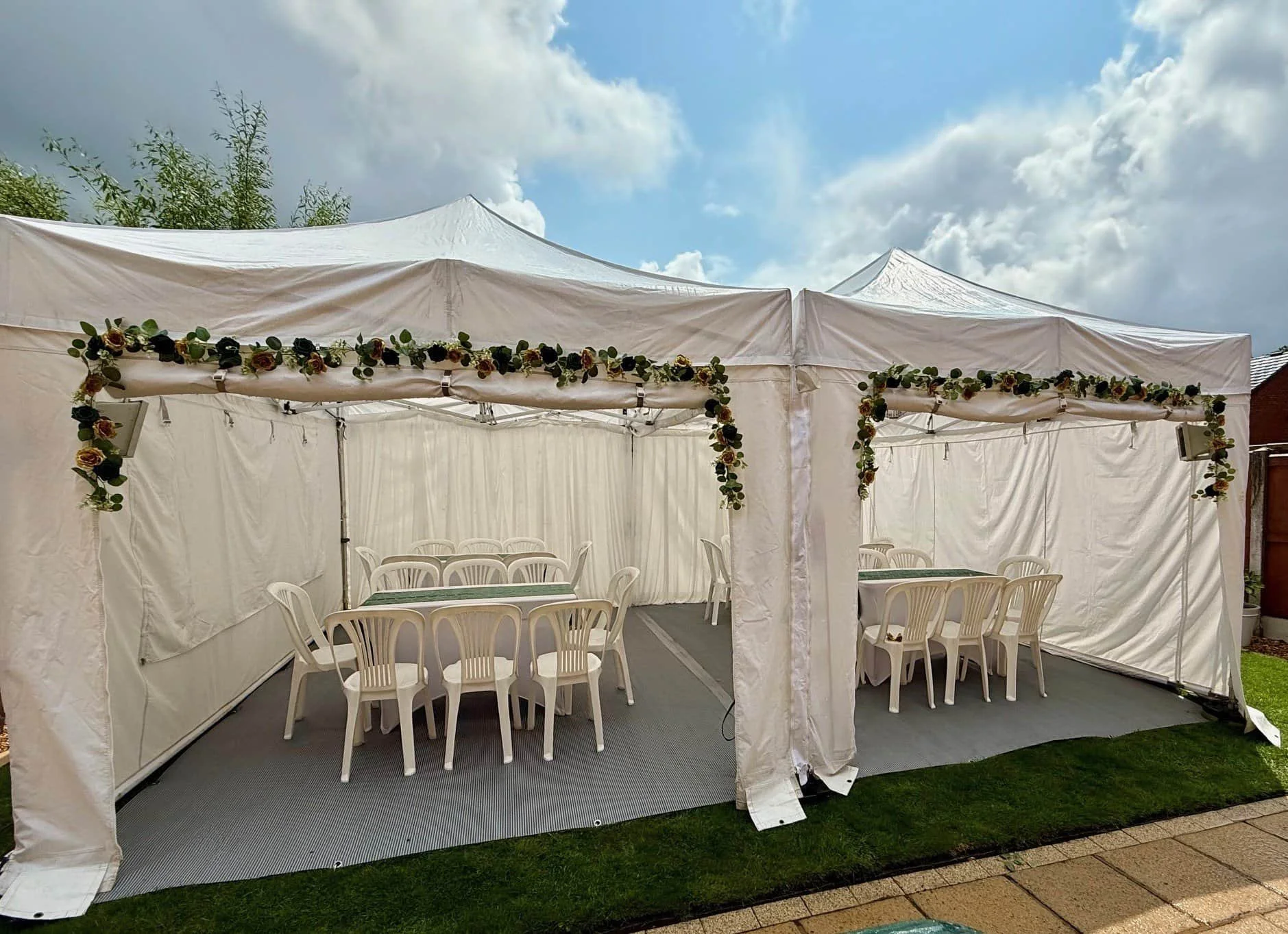 A white party tent decorated with a floral garland, set up outdoors with four tables and white plastic chairs inside.