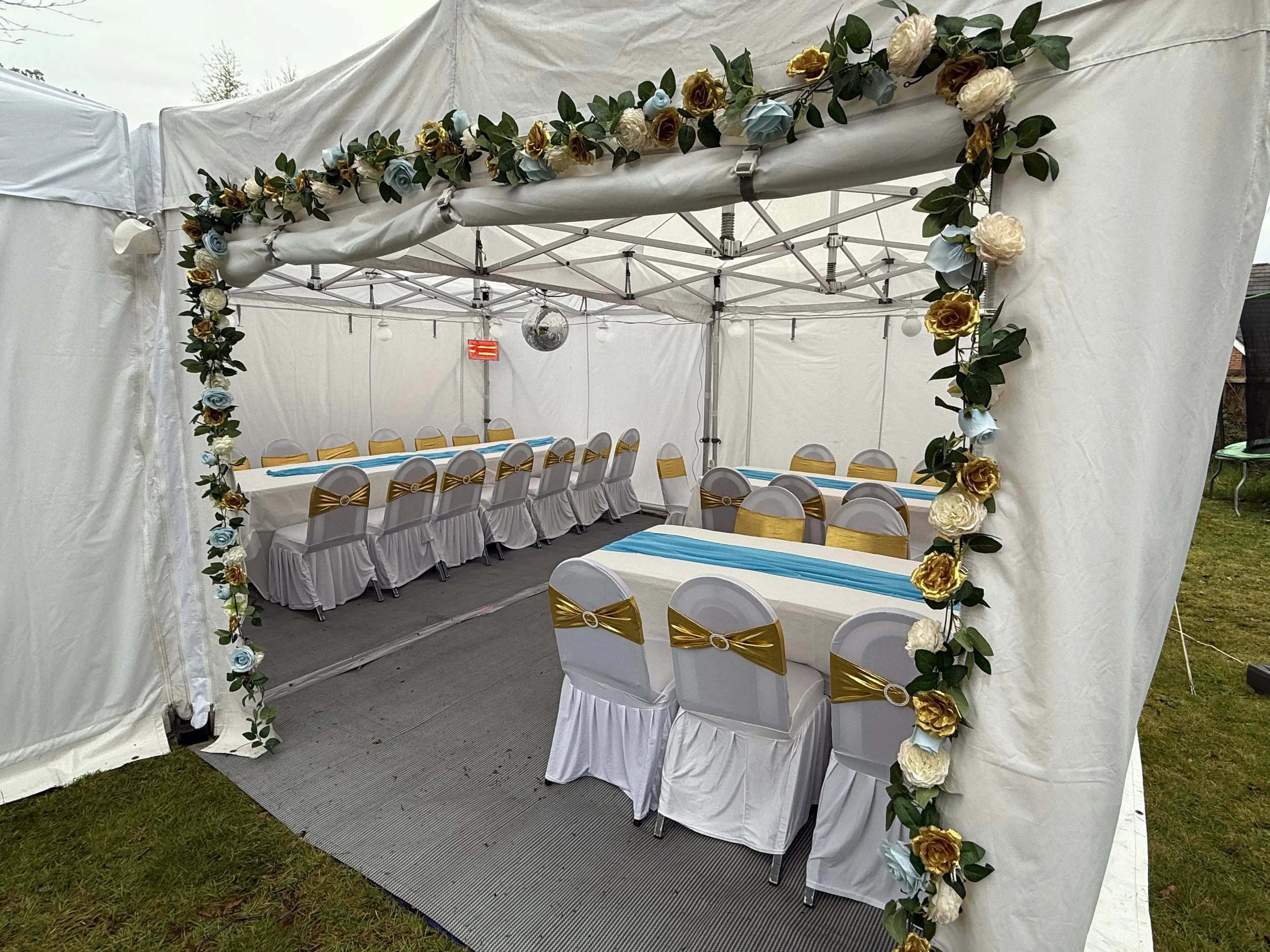 Decorated event tent with white drapes, floral garland, and set tables with white tablecloths, gold and blue accents, and chairs with gold sashes.