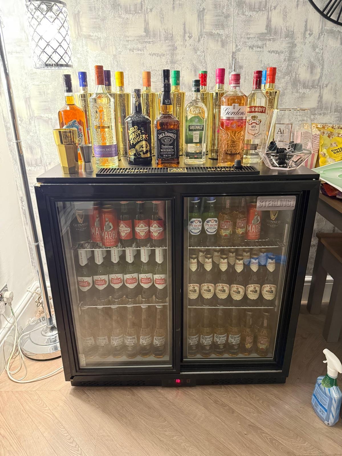 A mini bar refrigerator filled with bottled beers standing on a wood floor, with various liquor bottles and mixers on top, and a spray bottle on the right side.
