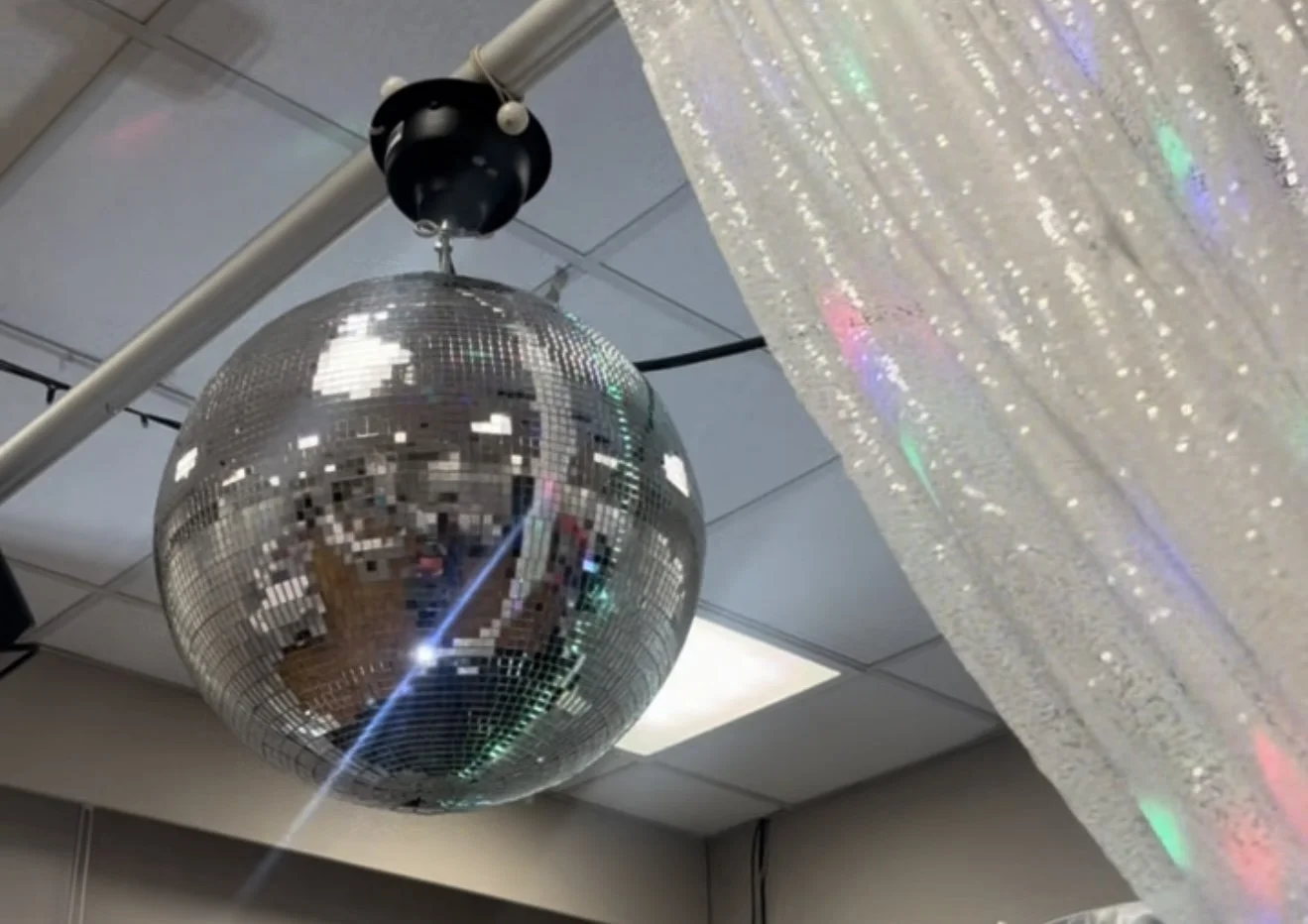 A large disco ball hanging from the ceiling reflects colorful lights onto a shiny curtain.