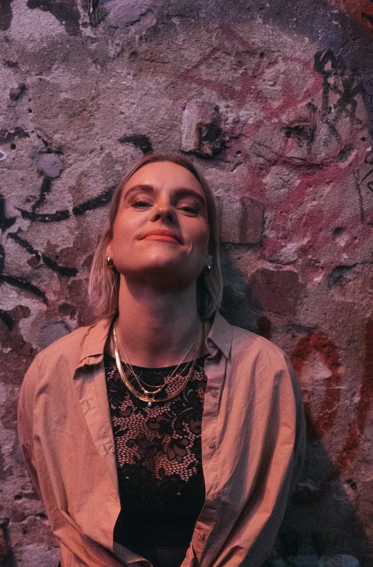Junge Frau mit Schulterlänge blondem Haar, trägt schwarze Spitzenbluse, beige Jacke und mehrere Goldketten, steht vor einer grauen, graffiti-besprühten Steinmauer im dunklen Raum.