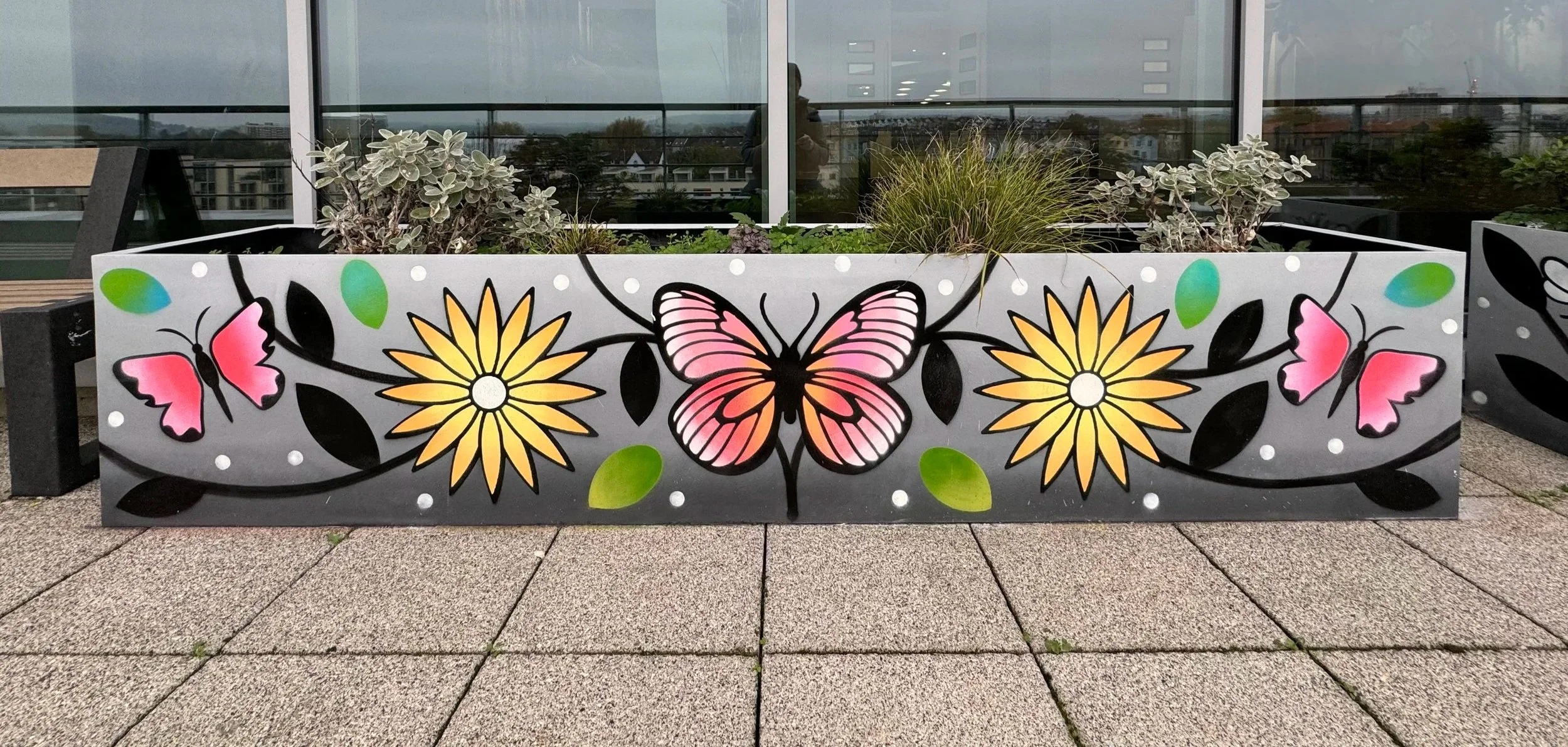Colorful outdoor planter with a pink butterfly, yellow flowers, a pink and black butterfly, green leaves, and white dots, located on a tiled balcony with a cityscape in the background.