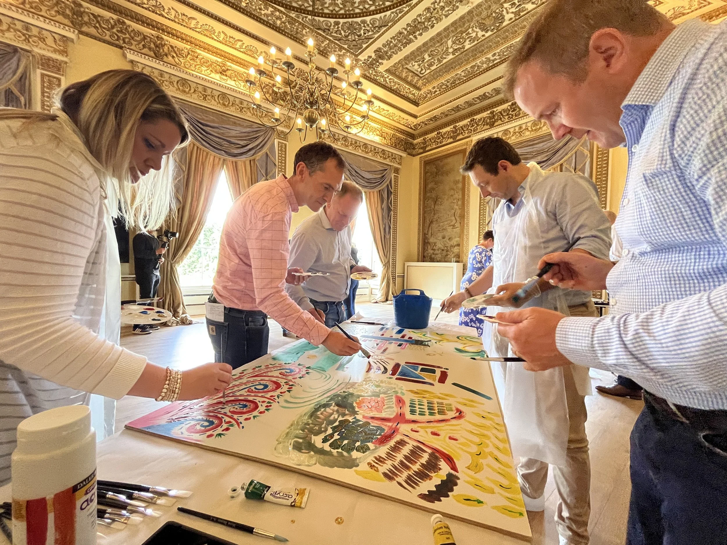 People painting a large colorful collaborative painting canvas in an ornate, well-lit room with gilded decorations and drapes.