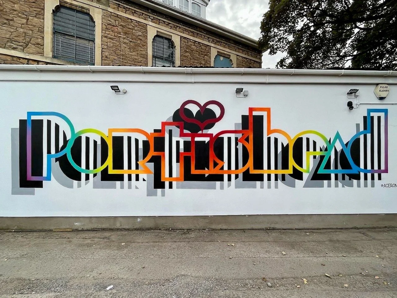 Colorful graffiti art on a white wall spelling out "Portland" with black and white striped letters, rainbow-colored outlines, and a black heart with red outline at the top center.
