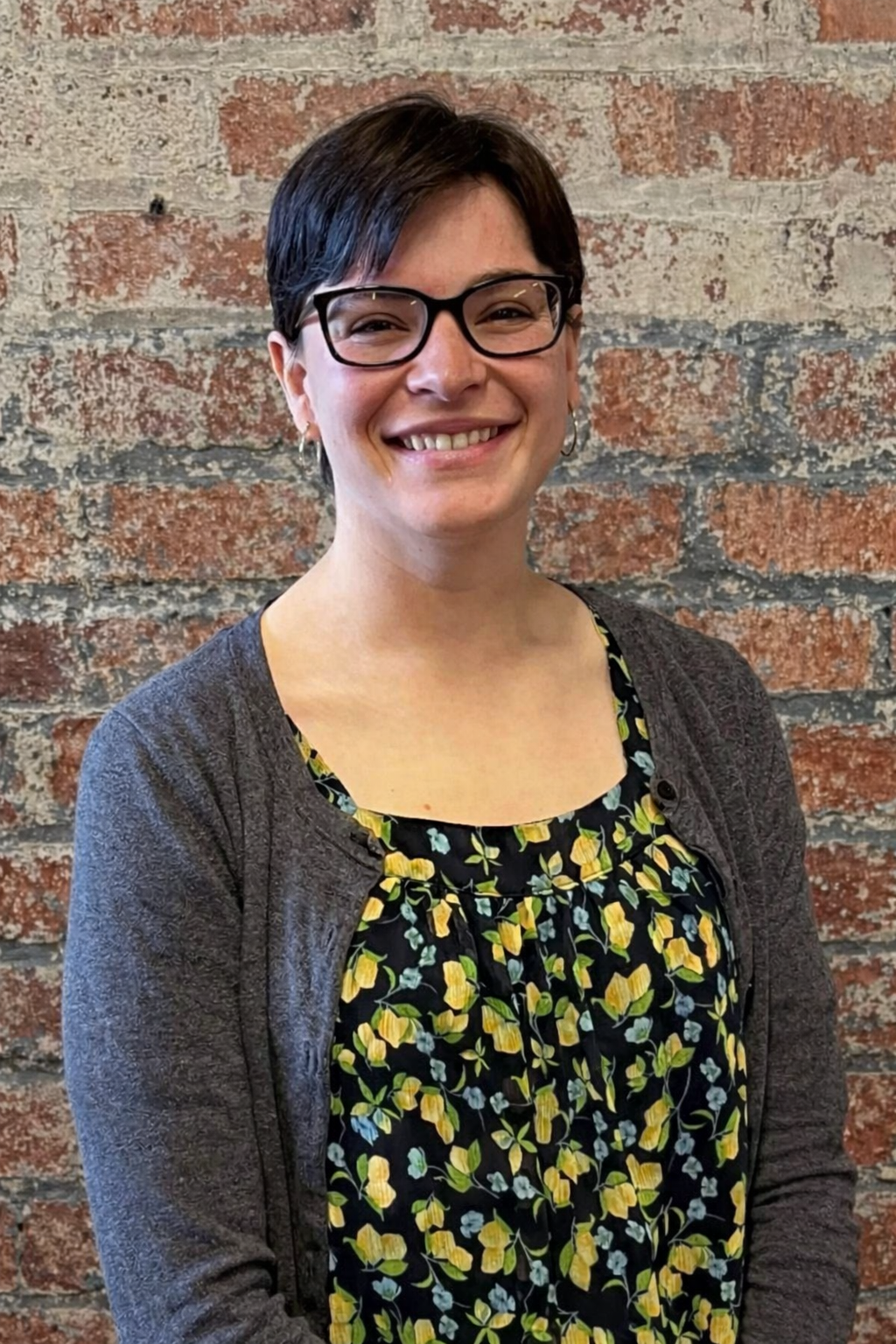 A woman with short dark hair and glasses smiling, wearing a floral top and a dark cardigan, standing against a brick background.