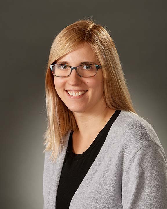 A woman with blonde hair, glasses, and a black top under a light gray cardigan smiling against a gray background.