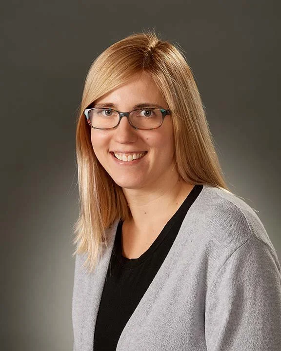 A woman with blonde hair, glasses, and a black top under a light gray cardigan smiling against a gray background.