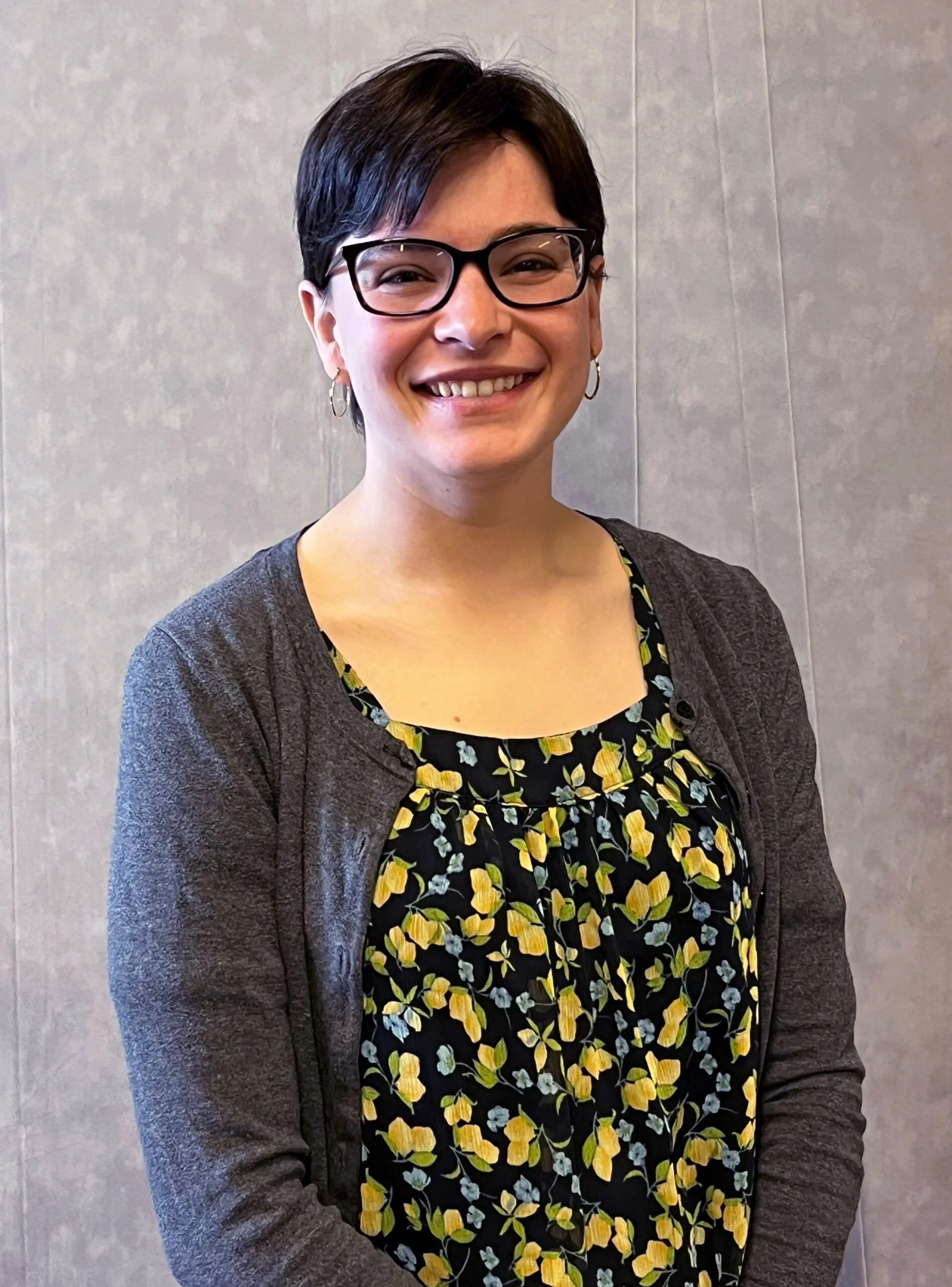 A woman with short dark hair and glasses smiling, wearing a floral top and a dark cardigan, standing against a neutral wall.