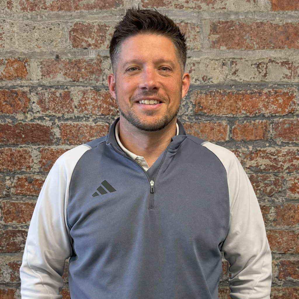 Picture of a man in a grey 1/4 zip shirt smiling against a brick background.
