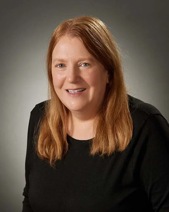 A woman with long red hair wearing a black top, smiling at the camera against a neutral gray background.