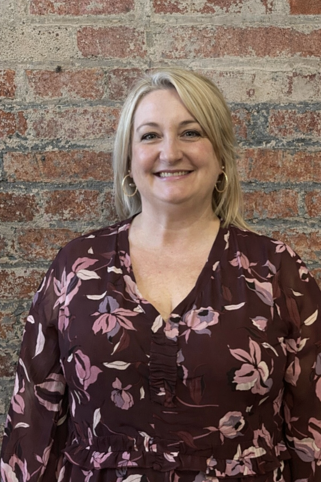 A woman with blonde hair smiling, wearing a dark floral blouse, standing against a neutral background.