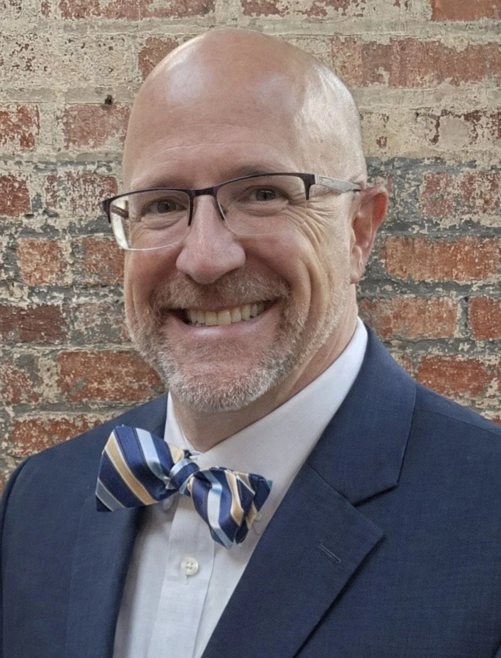 A man with glasses wearing a light blue shirt and striped bowtie smiling in front of a brick background