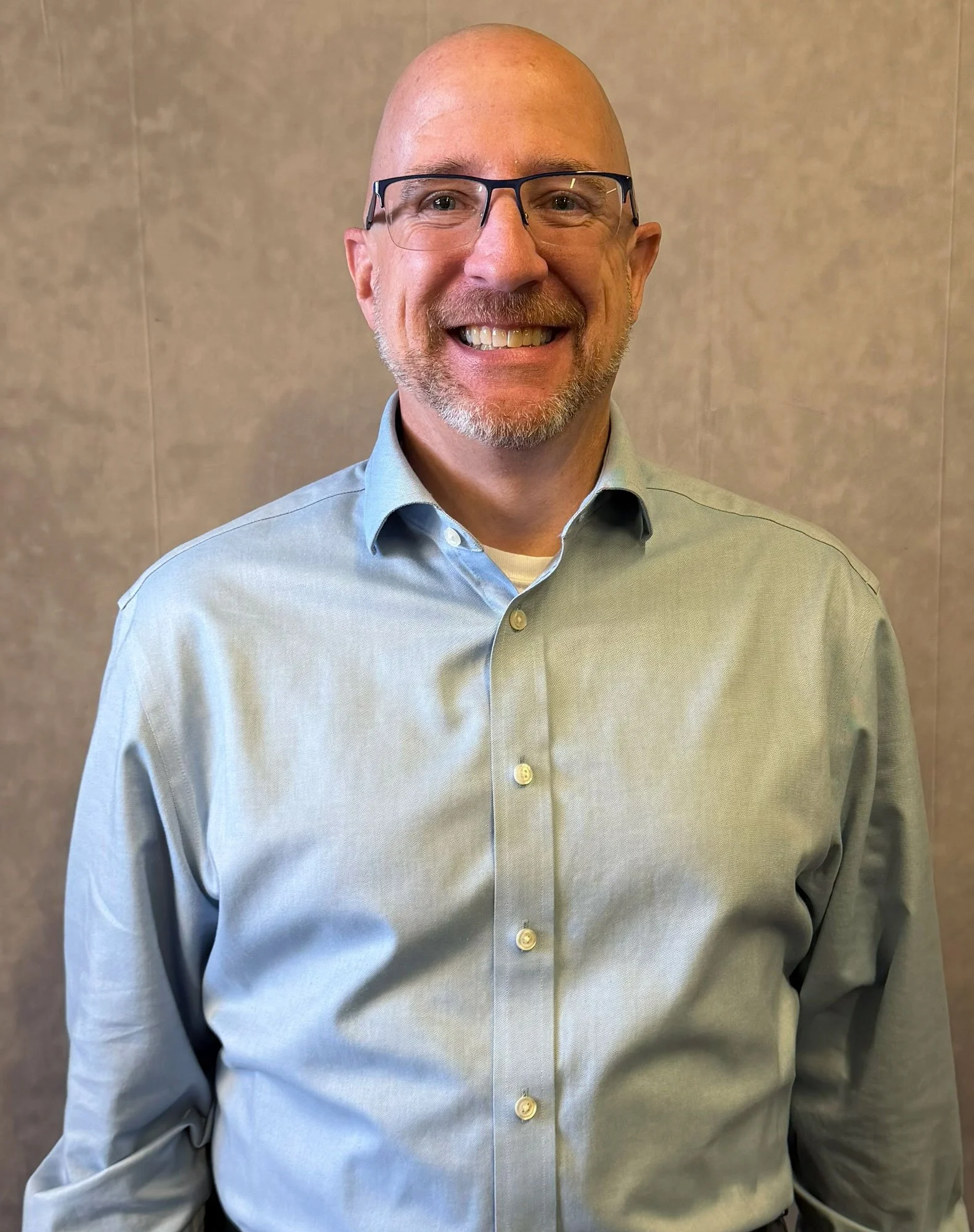 A man with glasses wearing a light blue checkered shirt smiling in front of a plain gray wall.