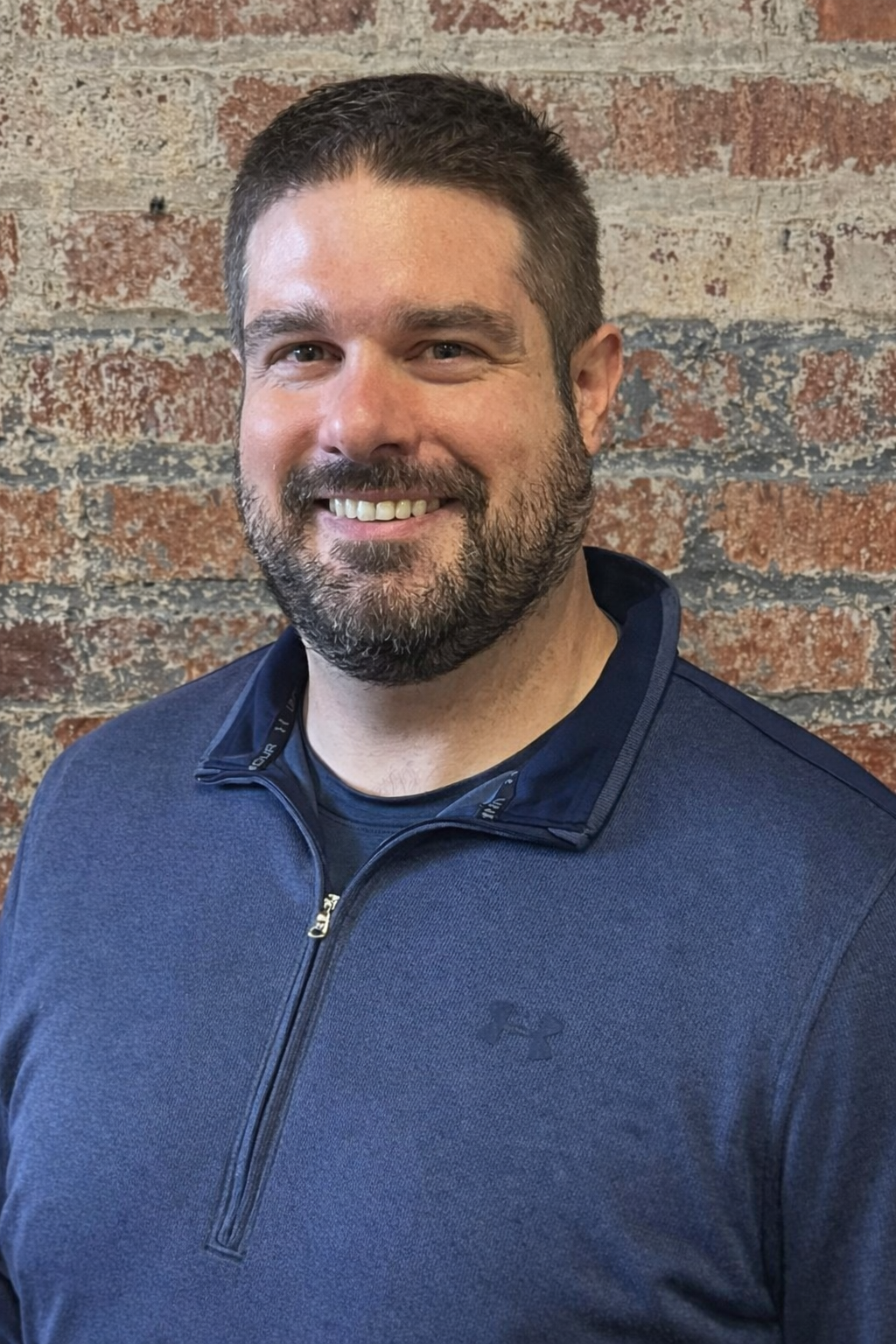 A smiling man with short dark hair and a beard, wearing a dark gray 1/4 zip shirt standing against a brick background..