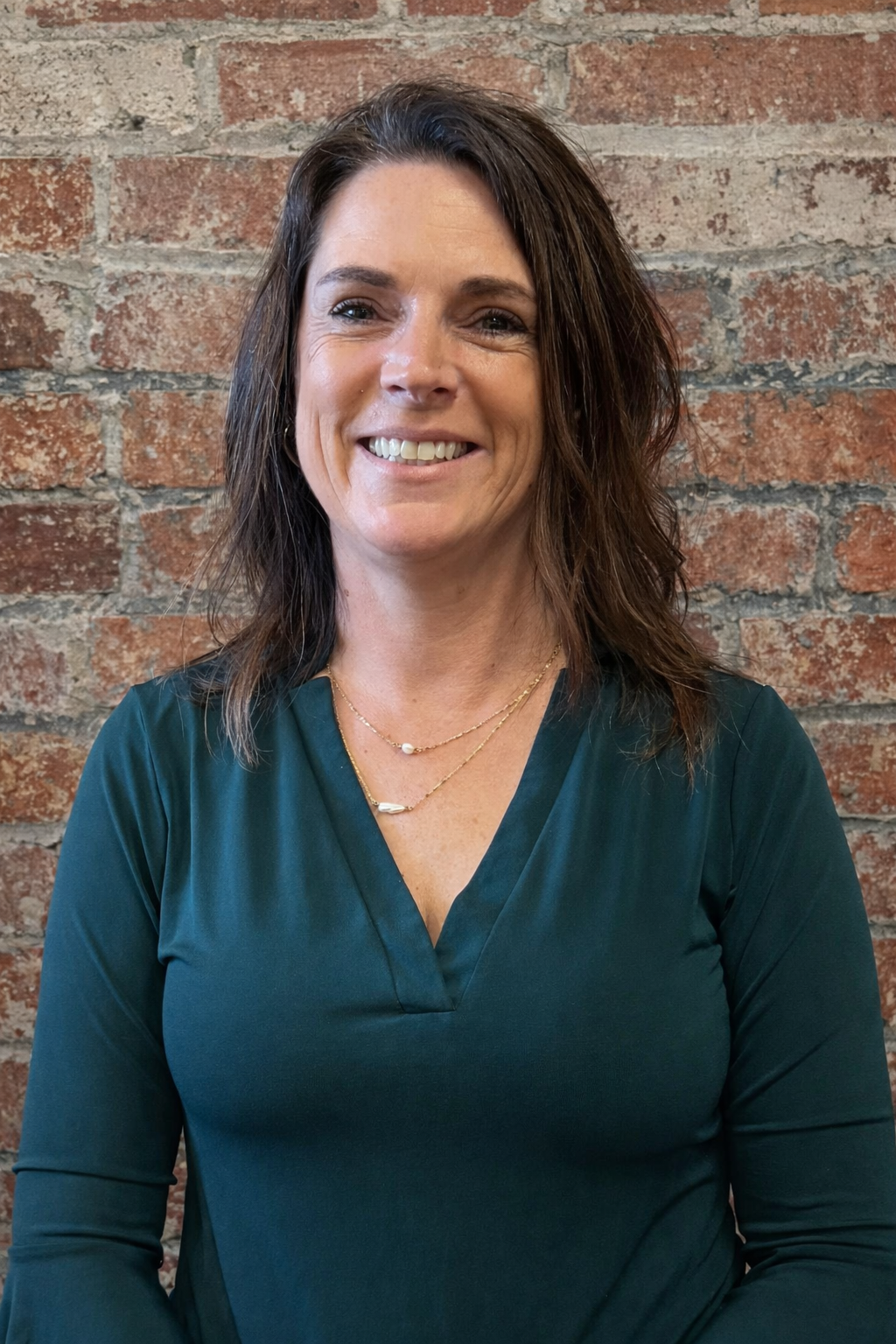 A woman with long brown hair, wearing a teal top, smiling against a brick background.