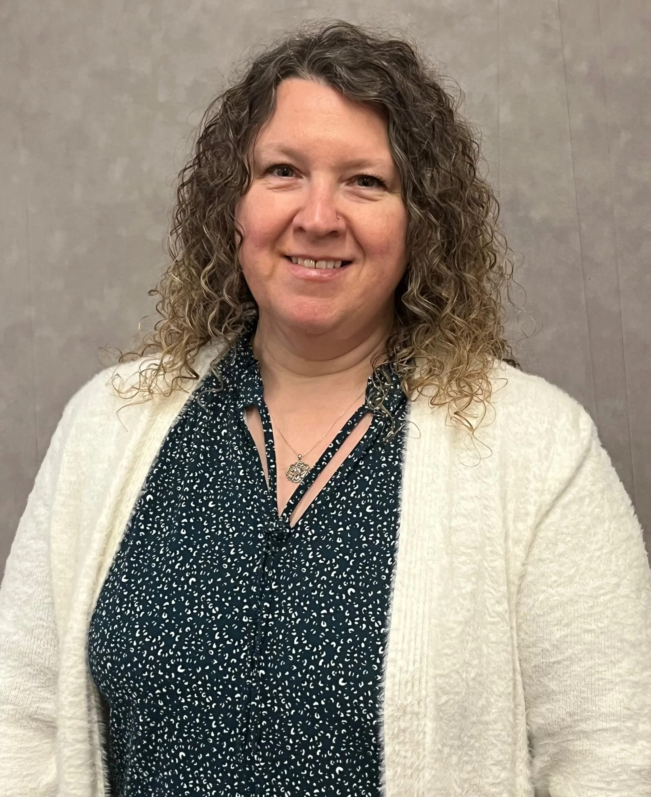 A woman with curly hair smiling, wearing a black and white patterned blouse, a cream cardigan, and a necklace, standing against a plain wall.