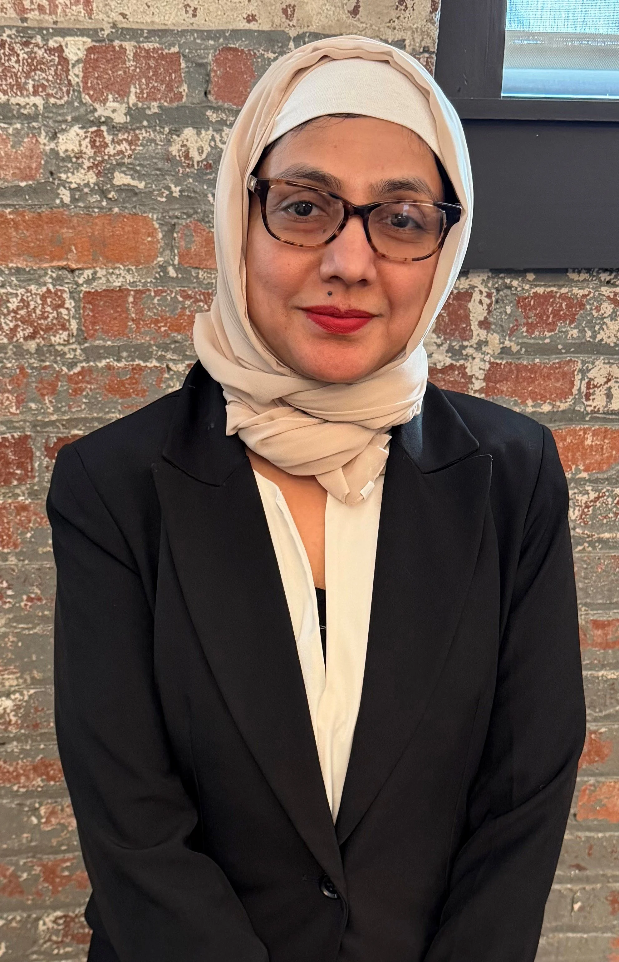 A woman in a tan headscarf and dark suit jacket against a brick background