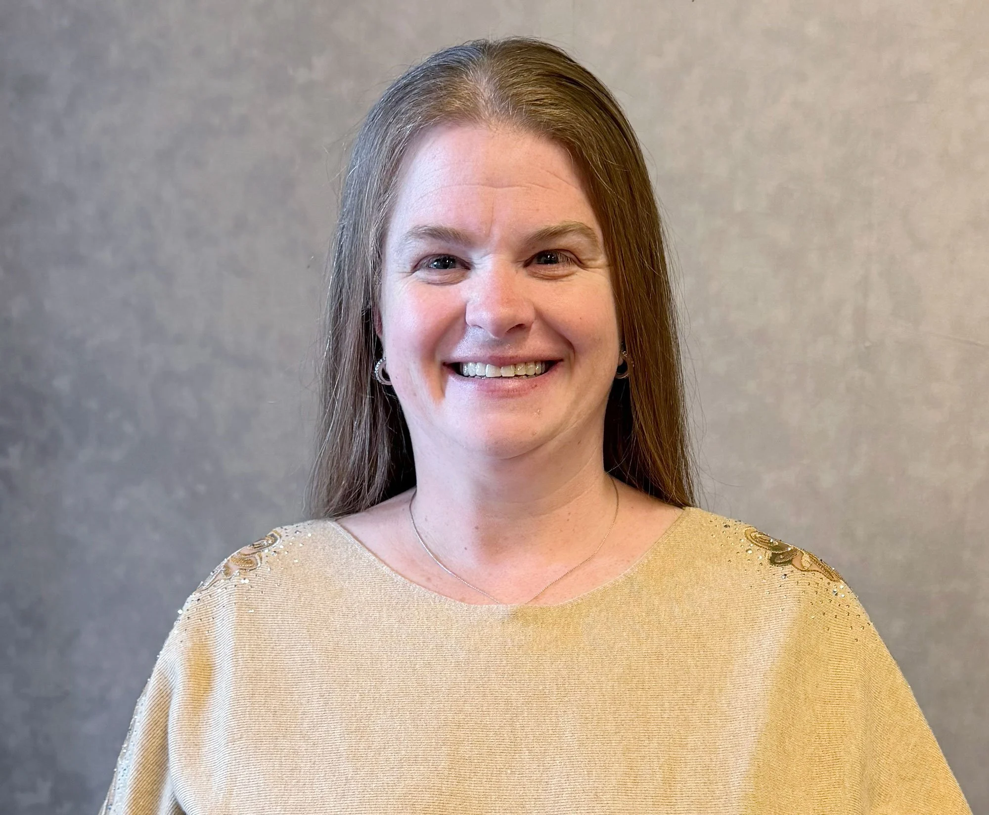 A woman with long brown hair, smiling, wearing a tan top against a neutral background.