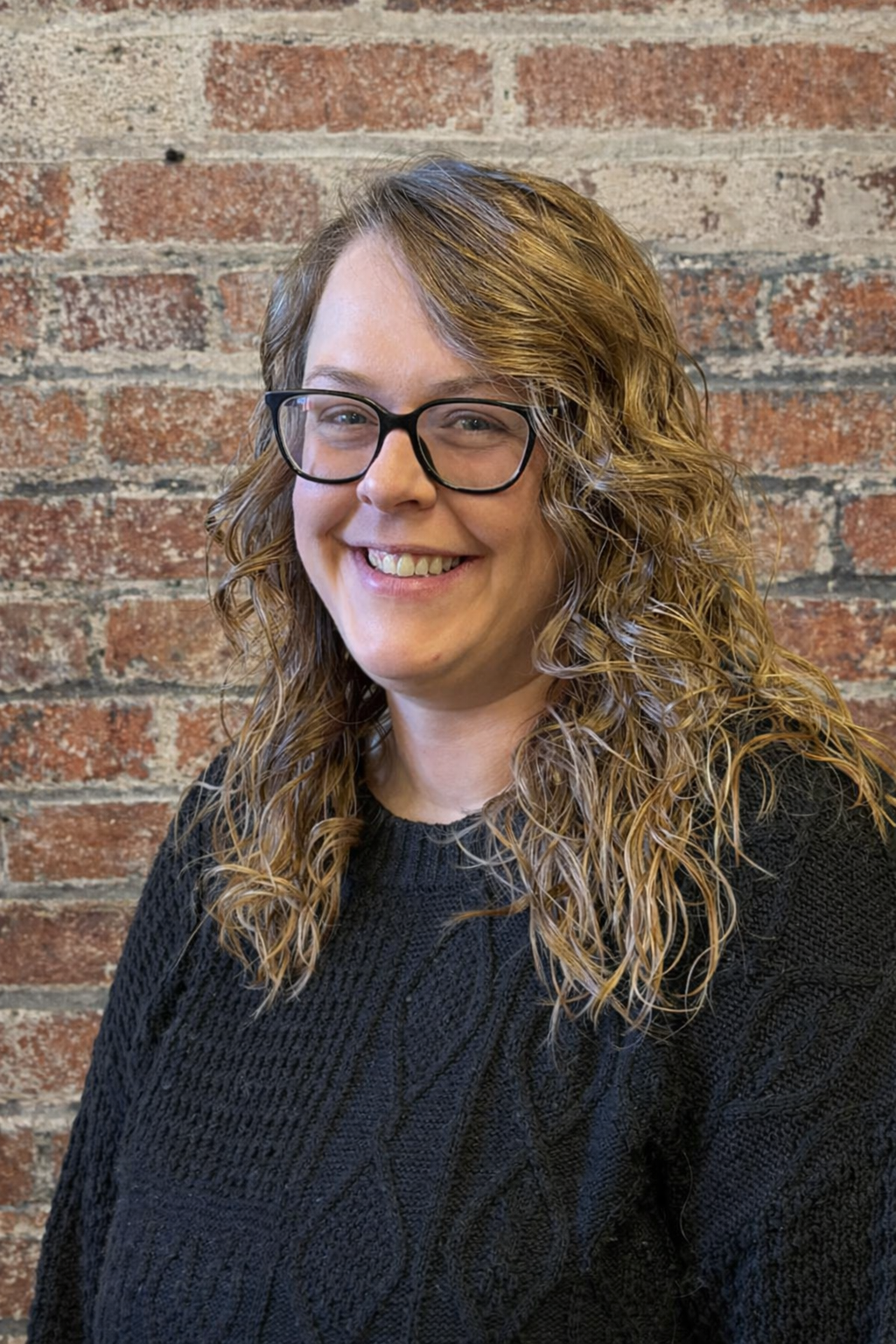 A woman with glasses and long, light brown hair wearing a dark sweater against a brick background