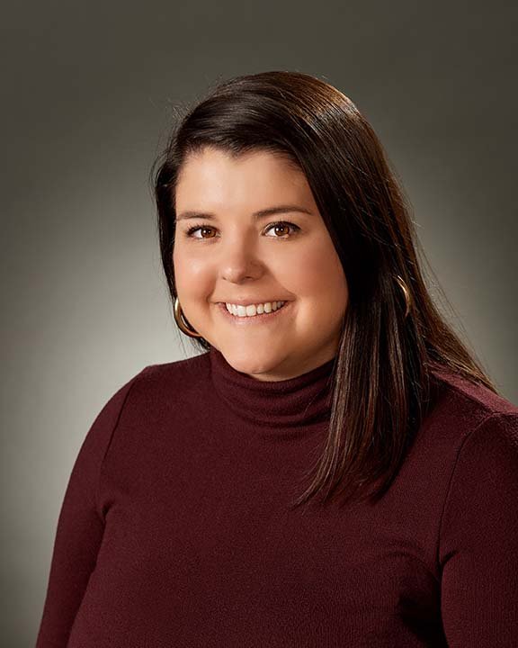 A woman with shoulder-length dark brown hair, wearing a maroon turtleneck sweater, smiling, against a gray gradient background.