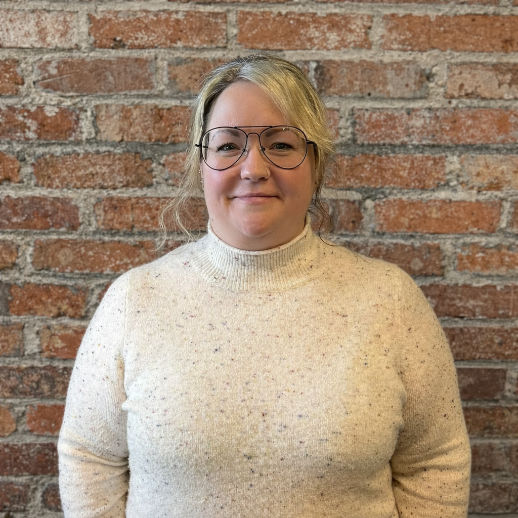 A woman with glasses and blonde hair, wearing a cream turtleneck sweater, standing against a brick background.
