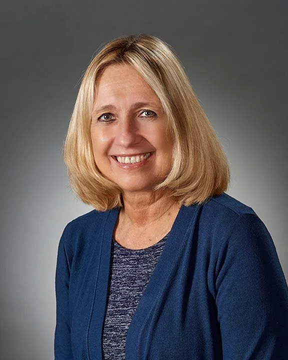 Portrait of a woman with blonde hair wearing a blue cardigan and a patterned top, smiling against a gray background.