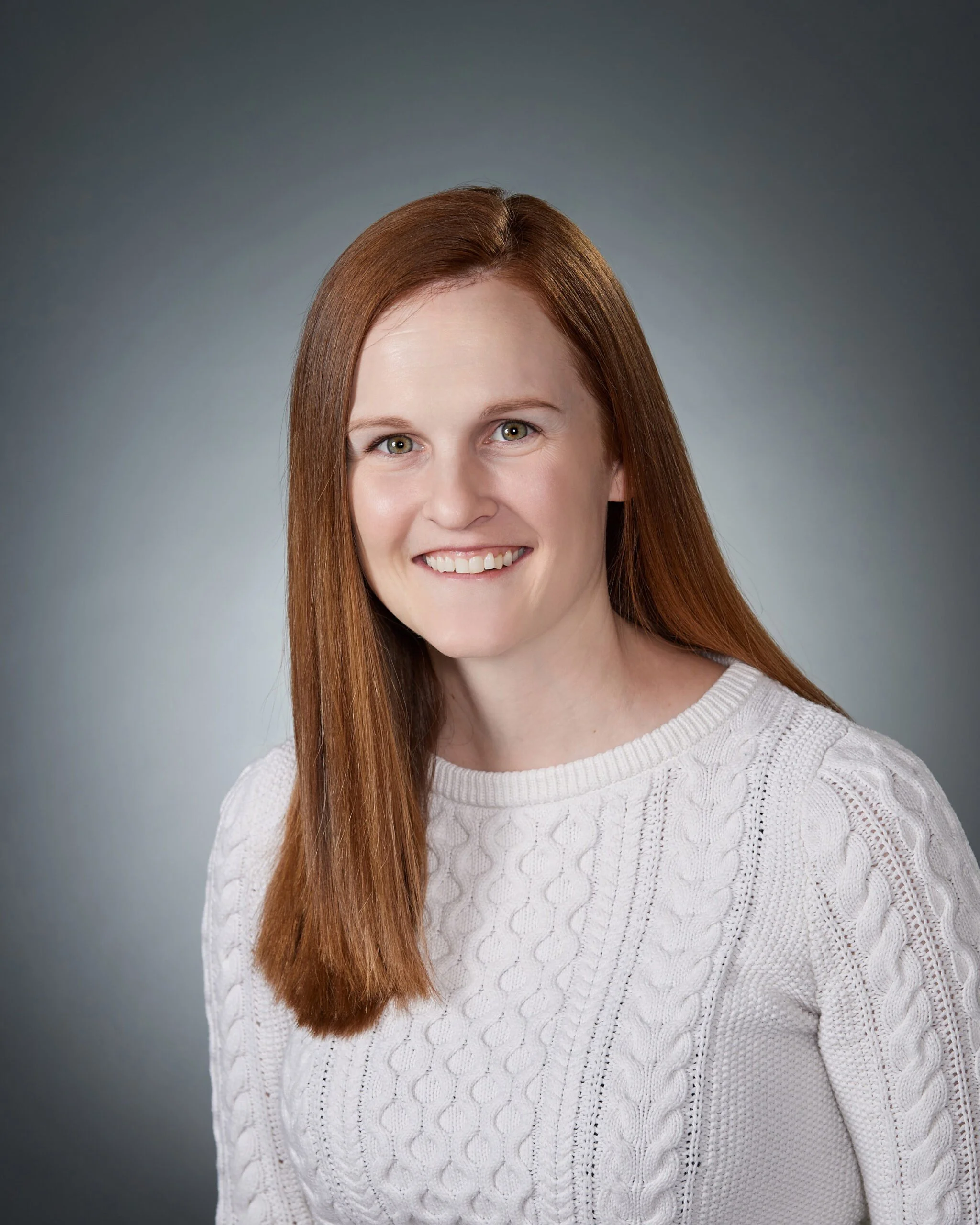 Headshot of a woman with long red hair smiling, wearing a white knitted sweater, against a gray background.