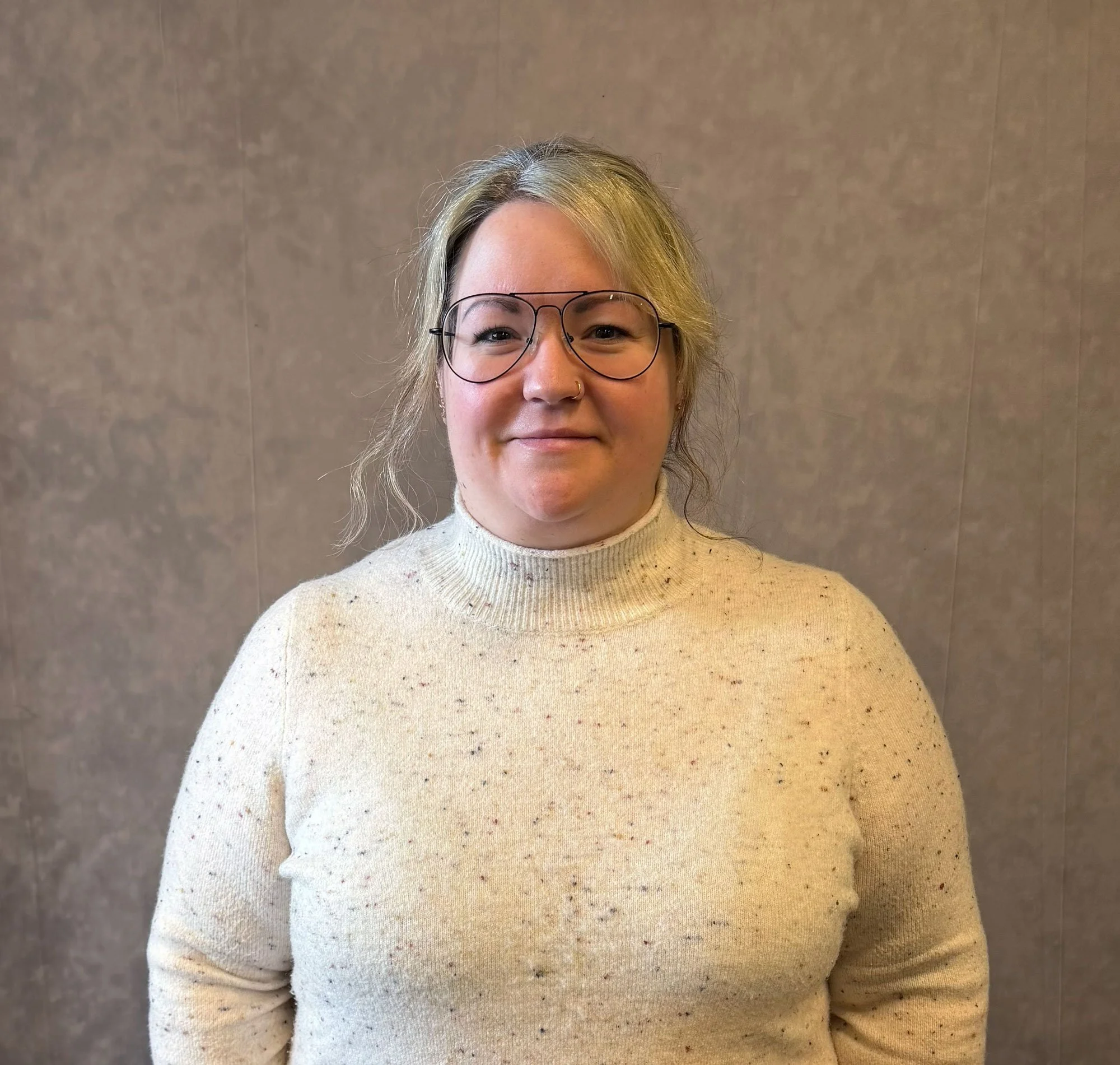 A woman with glasses and blonde hair, wearing a cream turtleneck sweater, standing against a plain background.