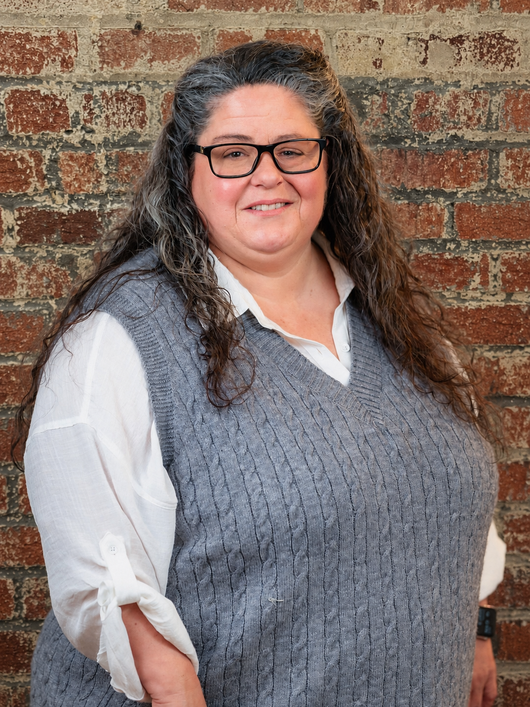 Photograph of a woman with long black hair, wearing glasses, a gray sweater, smiling in front of a brick background.