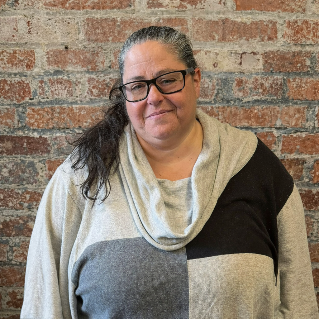 Photograph of a woman with long black hair, wearing glasses, a gray sweater, smiling in front of a brick background.