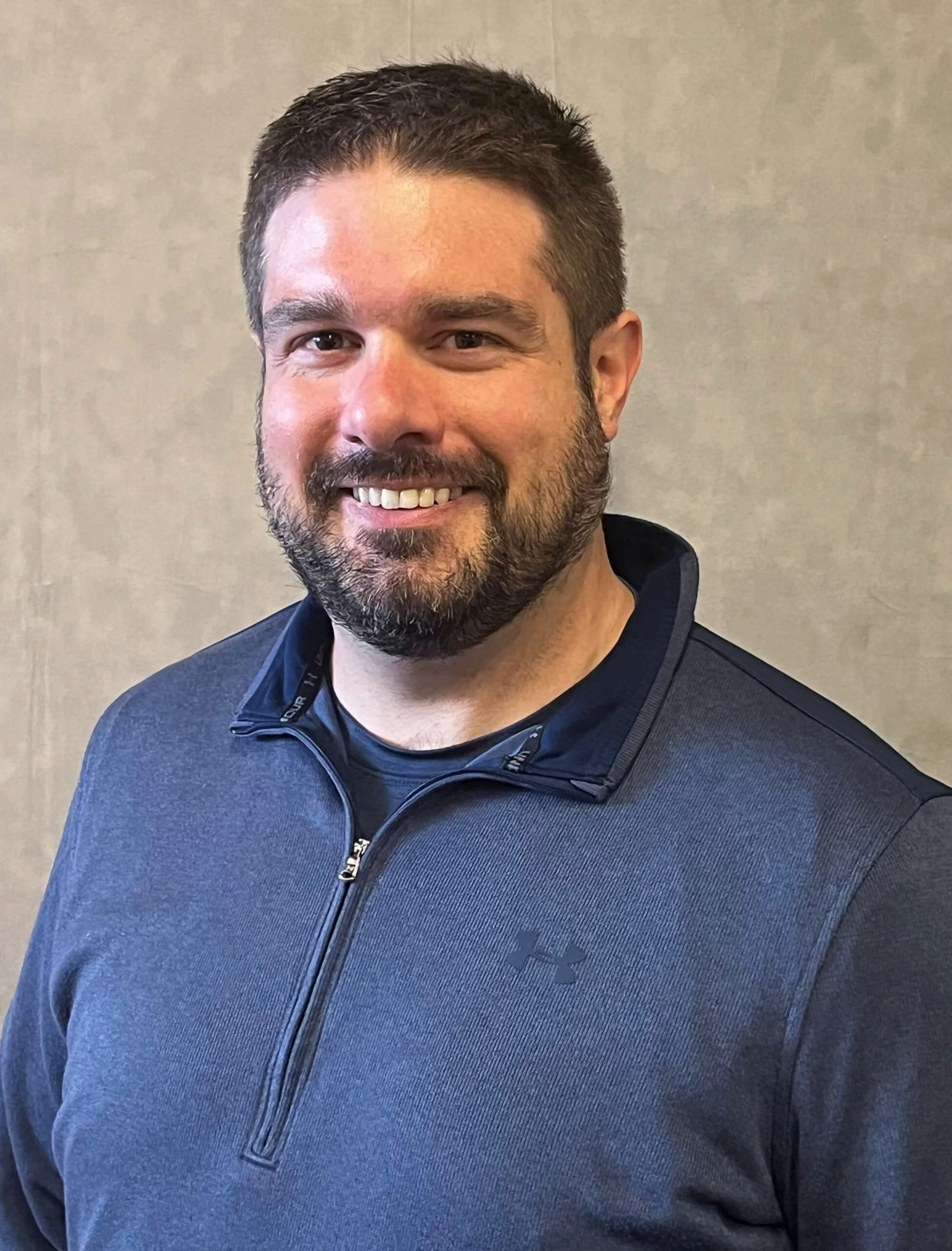 A smiling man with short dark hair and a beard, wearing a dark gray Under Armour jacket, standing against a plain beige wall.