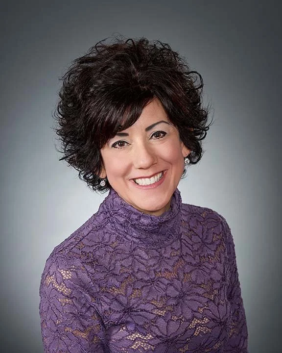 A woman with short, curly dark hair, smiling, wearing a purple lace top, against a gray background.