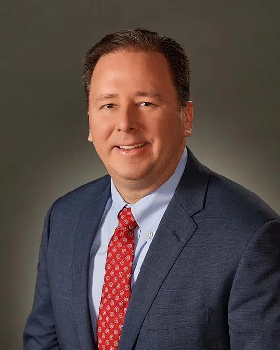 A smiling man in a blue suit, light blue dress shirt, and red patterned tie poses against a plain background.