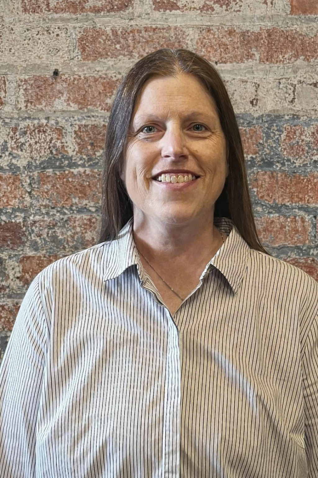 A woman with long dark hair smiling, wearing a striped button-up shirt, standing against a brick background.