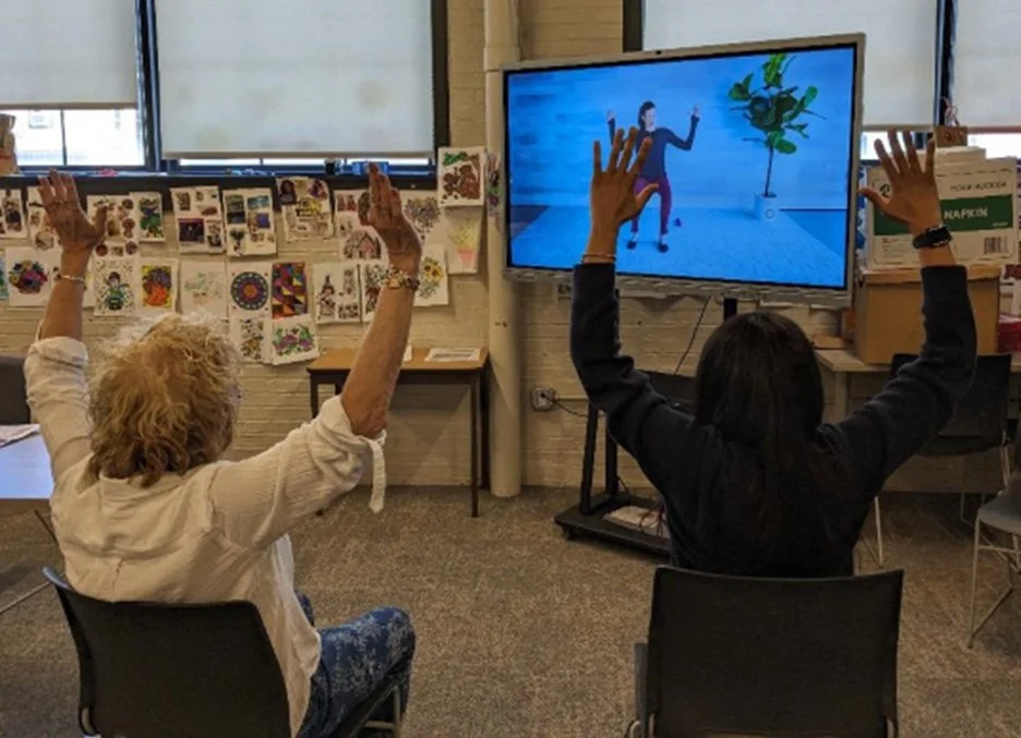 Two women sitting in chairs facing a television screen, participating in an interactive activity, with their hands raised towards the screen.
