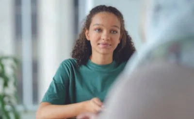 A young woman with curly hair wearing a teal shirt, sitting at a desk, looking at a computer screen.