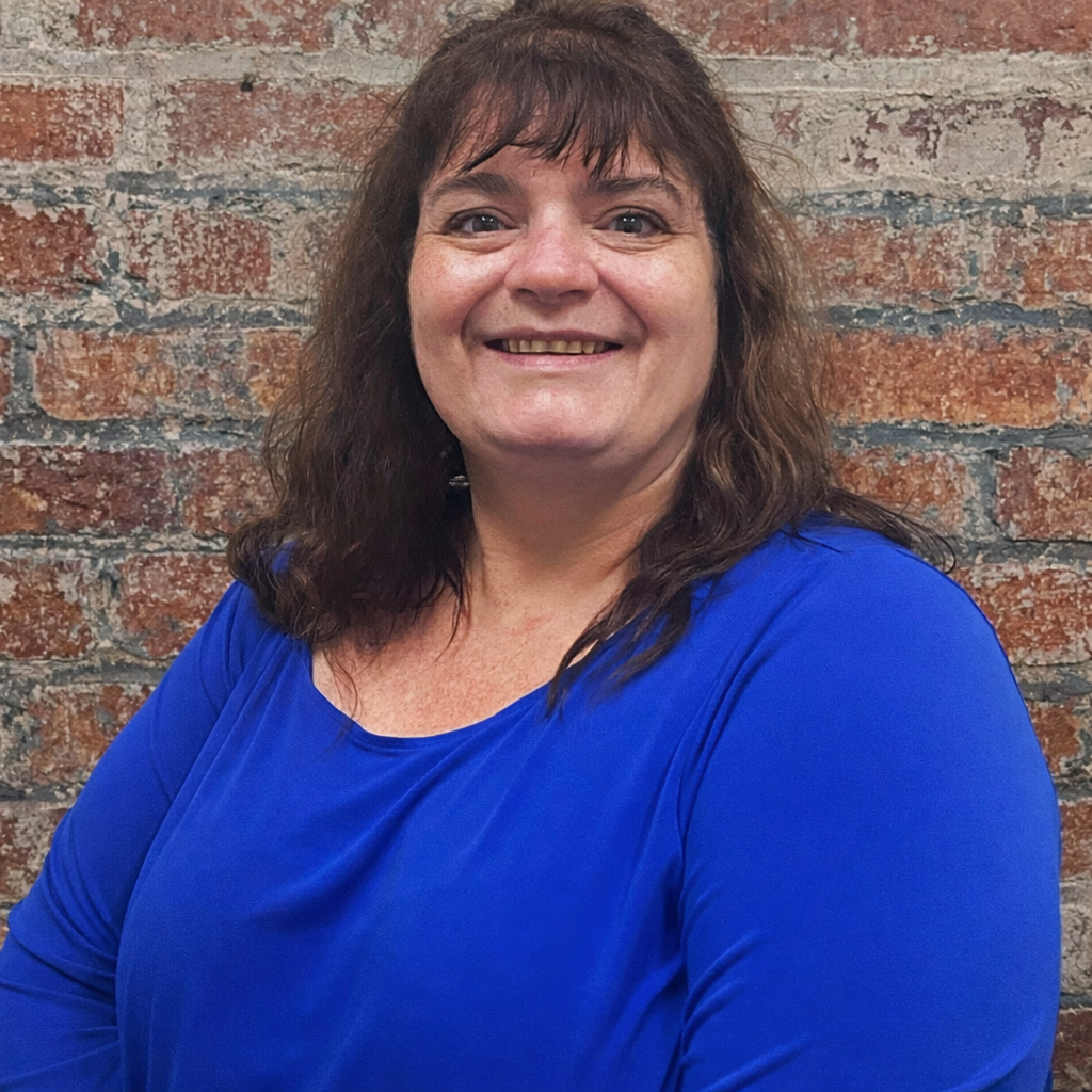 A woman with brown hair wearing a bright blue top, smiling in front of a brick background.