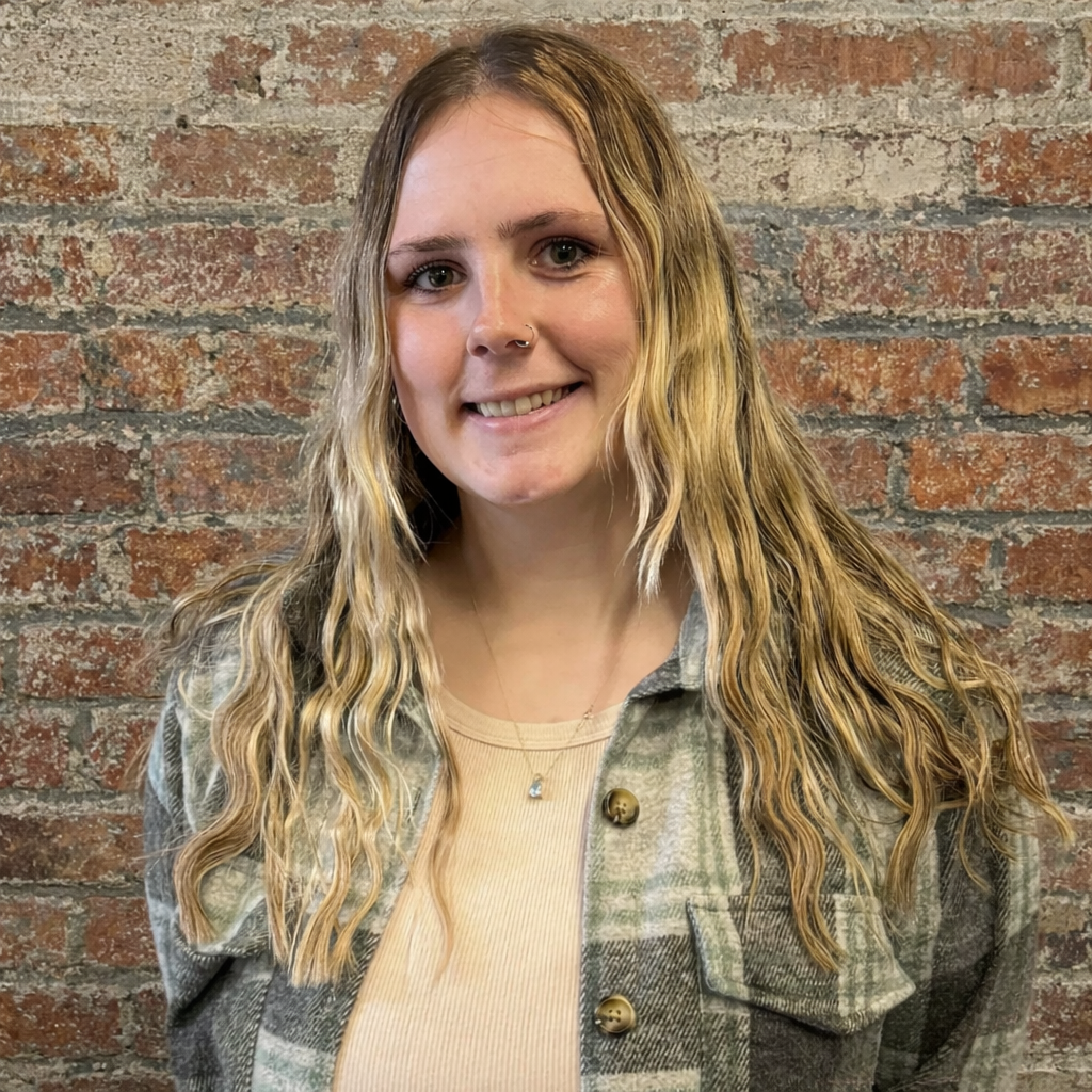 Picture of a woman with long blond hair in a plaid jacket against a brick background.