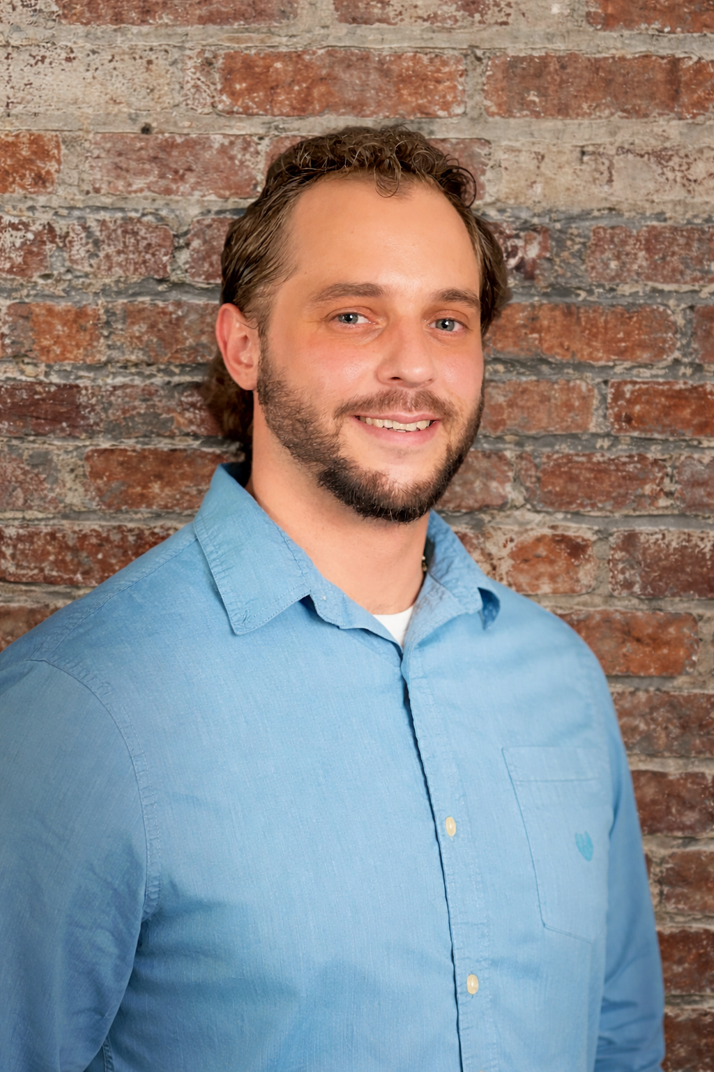 Picture of a man with a beard wearing a blue dress shirt in front of a brick background