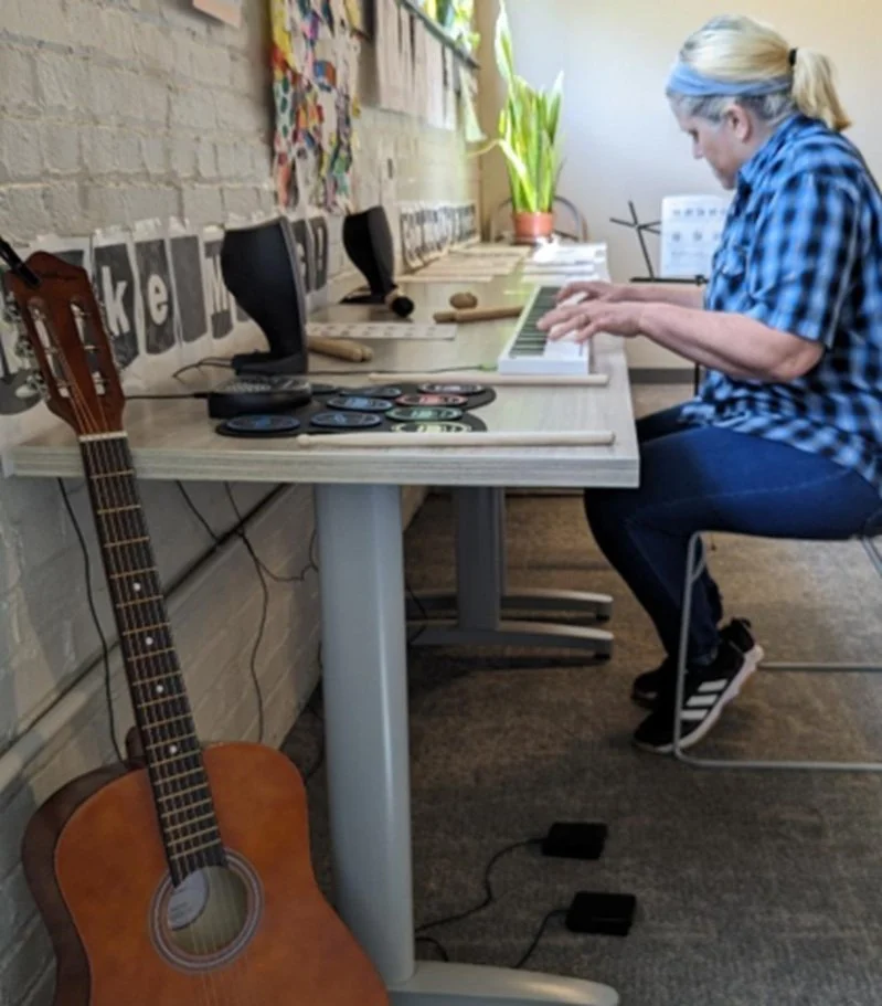 A woman sitting at a desk playing a small keyboard piano with an acoustic guitar in the foreground