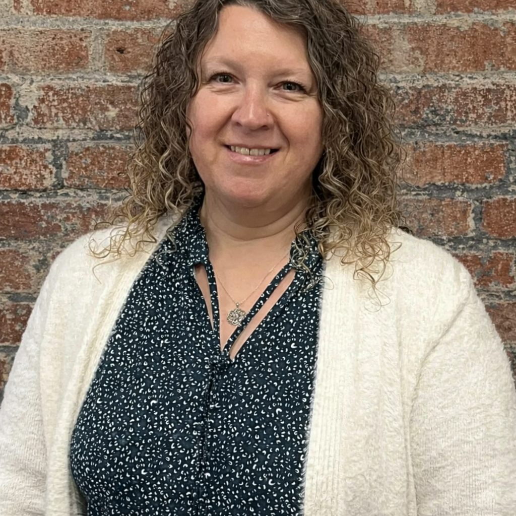 A woman with curly hair smiling, wearing a black and white patterned blouse, a cream cardigan, and a necklace, standing against a brick background..