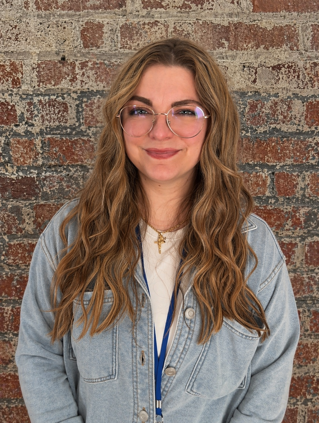 A woman with wavy hair, wearing a white top, a denim shirt and glasses, smiling at the camera against a brick background.