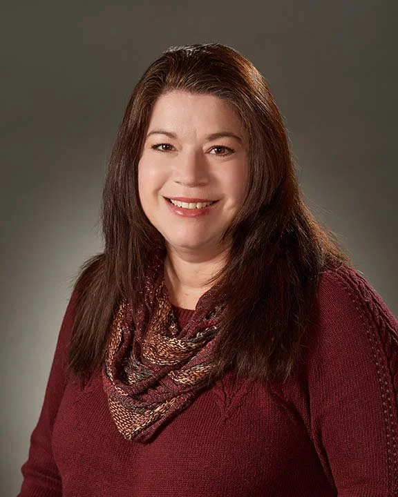 A woman with long brown hair, smiling, wearing a maroon sweater and a patterned scarf, against a neutral gray background.