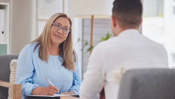 A woman with long blonde hair and glasses, wearing a light blue blouse, sits at a table and listens to a young man with short dark hair, wearing a white shirt, during a conversation in an office setting.